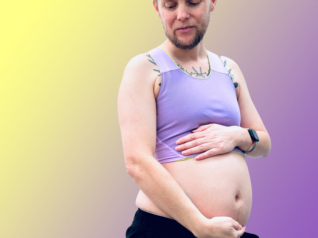 Andy, a non-binary trans masc person with a beard looks down at their pregnant belly with pride. They wear a lilac purple chest binder.